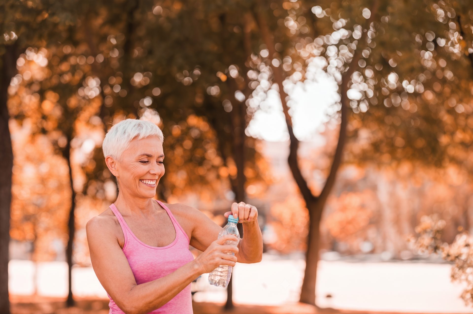 A smiling woman with short silver hair in a pink top opening a water bottle outdoors on an autumn day.